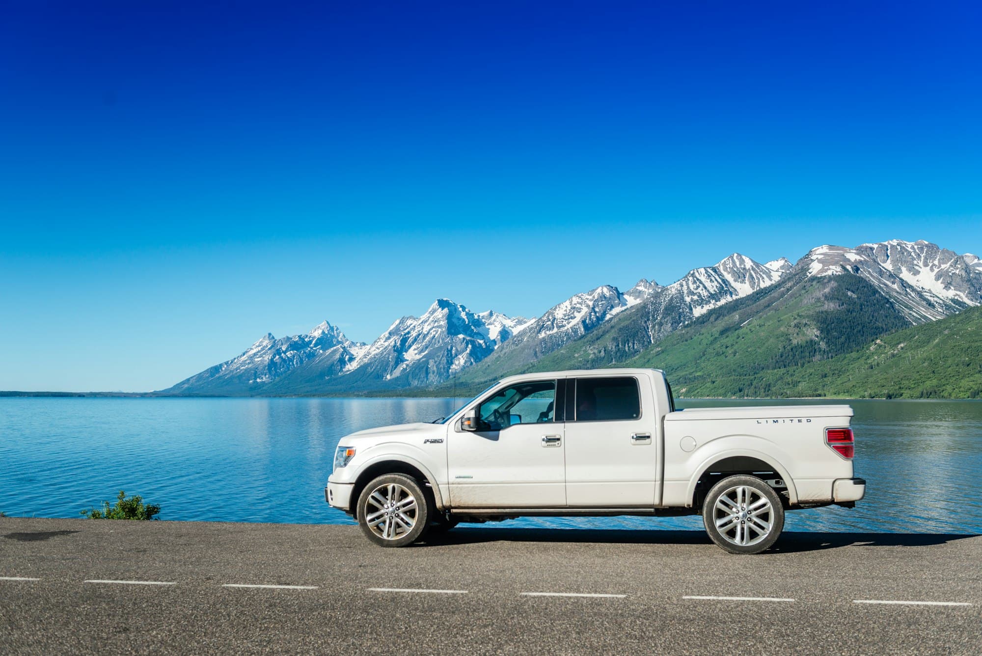 A white Ford F-150 SuperCrew parked on a lakeside road below snow-capped mountains